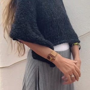 May include: A woman wearing a black knitted sweater with a grey pleated skirt. She is wearing a gold bracelet and a ring on her finger.