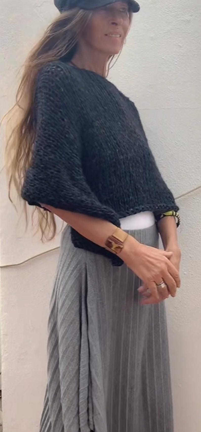 May include: A woman wearing a black knitted sweater with a grey pleated skirt. She is wearing a gold bracelet and a ring on her finger.