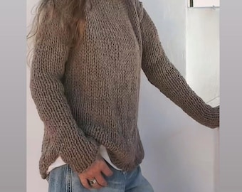 May include: A person wearing a dark blue cap, a brown knit sweater, and light blue jeans. The sweater has a crew neck and long sleeves. The jeans are loose-fitting. The person is leaning against a white wall.