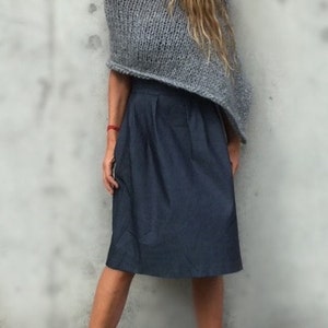 May include: A woman wearing a grey knitted poncho and a dark blue skirt. She is standing in front of a grey wall and wearing purple sandals.
