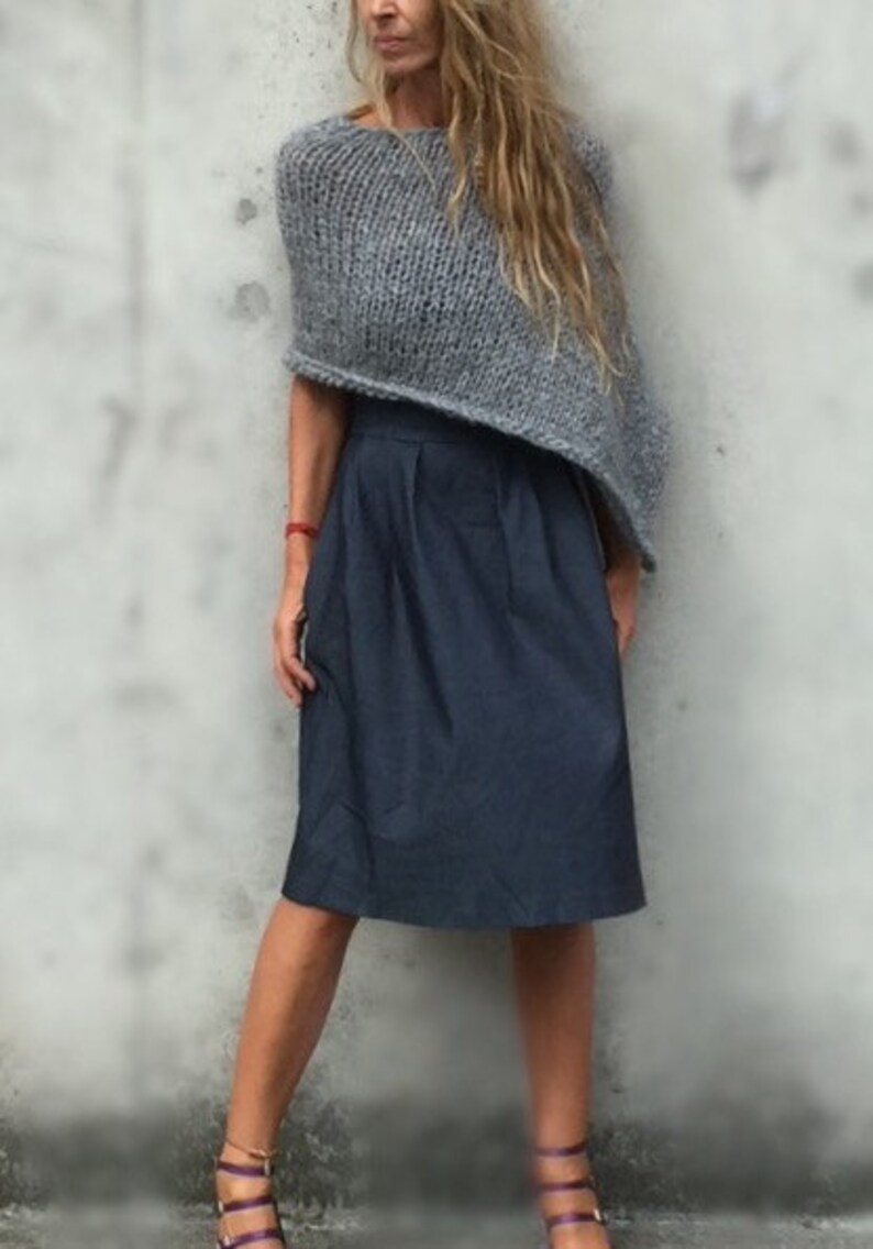 May include: A woman wearing a grey knitted poncho and a dark blue skirt. She is standing in front of a grey wall and wearing purple sandals.