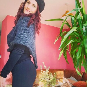 May include: A woman wearing a black beret and a grey knitted shawl over a black long-sleeved shirt and black trousers. She is standing in a room with a pink wall and a large green plant in the background.