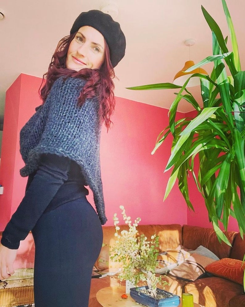 May include: A woman wearing a black beret and a grey knitted shawl over a black long-sleeved shirt and black trousers. She is standing in a room with a pink wall and a large green plant in the background.