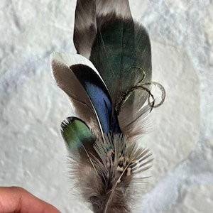 Op de afbeelding: Een decoratieve compositie van veren in bruin, groen en blauw, bevestigd aan een messing patroonhuls. De veren zijn in een cluster gerangschikt. De tekst "SciannaArt By Yumi" is zichtbaar.