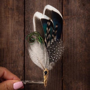 May include: A decorative feather hair clip featuring a gold-toned metal base with a black gemstone and an arrangement of feathers in shades of white, gray, teal, and black. The clip is held by a silver-toned metal clip. The text "SCIANNAARTBYYUMI" is at the bottom.