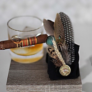 May include: A glass of whiskey with ice and a cigar resting on the rim of the glass. A black fabric with a shotgun shell and feathers is next to the glass on a wooden surface.
