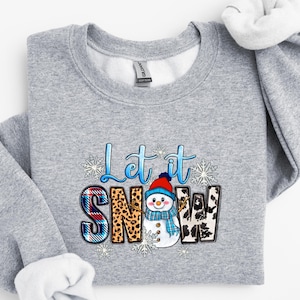 Op de afbeelding: Een grijs sweatshirt met een feestelijk ontwerp met een sneeuwpop en de tekst "Let it Snow". De letters zijn versierd met ruitjes, luipaardprint en andere patronen.