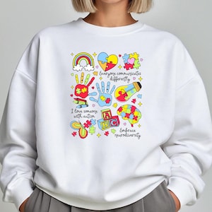 May include: White sweatshirt with a colorful graphic design featuring puzzle pieces, a rainbow, a lightbulb, a handprint, and the text "Everyone communicates differently", "I love someone with autism", "ABC", and "Embrace neurodiversity".