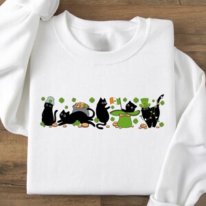May include: White sweatshirt with a black cat design. The cats are wearing green hats and holding shamrocks and gold coins. The text "Happy St. Patrick's Day" is printed on the sweatshirt.