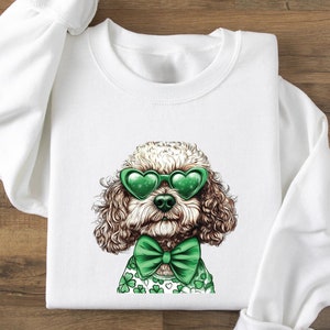 May include: White sweatshirt with a brown and white dog wearing green heart-shaped sunglasses and a green bow tie. The dog is wearing a green shirt with shamrock designs.