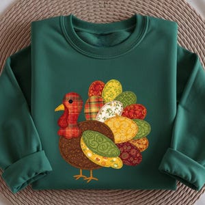 May include: A dark green sweatshirt featuring a colorful turkey design. The turkey has a red plaid head and a brown body, with a tail made of various patterned fabric ovals in shades of yellow, green, and red. The sweatshirt is folded on a woven mat.
