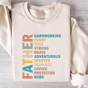 May include: A beige sweatshirt with the word "Father" spelled vertically in colorful letters. The letters are outlined in yellow and have a dotted pattern. The words "Hardworking, Funny, Wise, Strong, Brave, Adventurous, Devoted, Fearless, Loving, Protector, Hero" are listed vertically below the word "Father".