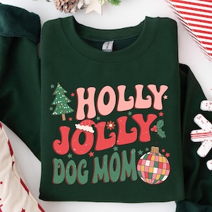May include: Green Christmas sweatshirt with the text "Holly Jolly Dog Mom" in a retro font. The design features a Christmas tree, a Santa hat, a holly sprig, and a disco ball ornament.