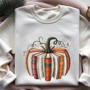 May include: A cream-colored crewneck sweatshirt featuring a central graphic of a pumpkin constructed from open, upright books. The books have varied orange, red, and green spines, with a dark green, twisted stem emerging from the top center.