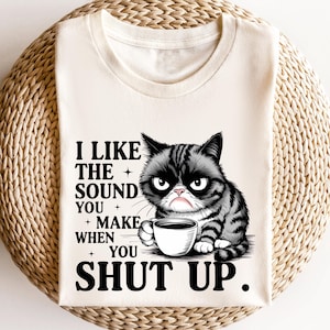 May include: Cream-colored t-shirt featuring a grumpy cat illustration with a coffee cup. The text reads, "I LIKE THE SOUND YOU MAKE WHEN YOU SHUT UP." The cat is black and gray with a displeased expression.