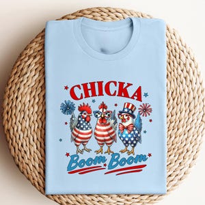 May include: Light blue t-shirt featuring a graphic of three cartoon chickens dressed in patriotic attire. The text "Chicka Boom Boom" is displayed in red and blue. The chickens are decorated with the American flag.