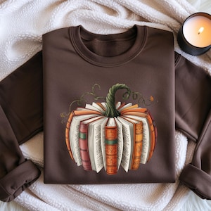 May include: A dark brown crewneck sweatshirt featuring a graphic of a pumpkin constructed from open books with colorful spines and a green stem. The design is centered on the chest, showcasing various book covers in shades of orange, red, and green.