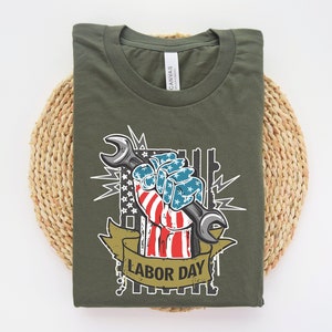 May include: A green t-shirt with a graphic of a hand holding a wrench. The hand is painted with the American flag and the text "Labor Day" is below the graphic.