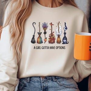 May include: Cream-colored sweatshirt featuring a row of whimsical broom designs in various colors and styles. The phrase "A GIRL GOTTA HAVE OPTIONS" is printed below the brooms. The person is holding an orange mug.