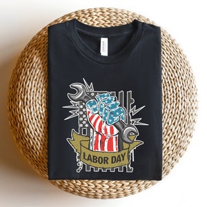 May include: Black t-shirt with a graphic of a hand holding a wrench. The hand is painted in the colors of the American flag and the wrench is silver. The graphic is surrounded by stars and the text "Labor Day".