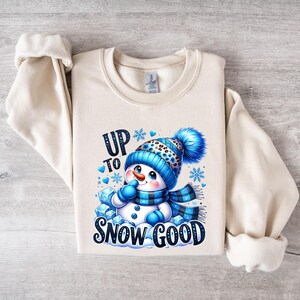 Peut inclure: Un sweat-shirt cr&egrave;me avec un motif de bonhomme de neige bleu et blanc. Le bonhomme de neige porte un chapeau bleu et blanc avec un imprim&eacute; l&eacute;opard noir et blanc. Le texte "Up to Snow Good" est imprim&eacute; en noir sous le bonhomme de neige.
