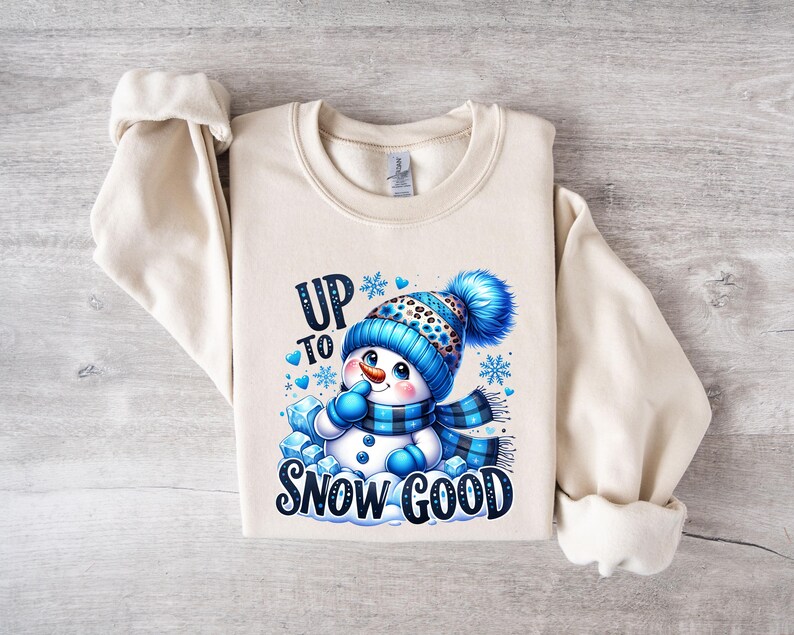 Peut inclure: Un sweat-shirt cr&egrave;me avec un motif de bonhomme de neige bleu et blanc. Le bonhomme de neige porte un chapeau bleu et blanc avec un imprim&eacute; l&eacute;opard noir et blanc. Le texte "Up to Snow Good" est imprim&eacute; en noir sous le bonhomme de neige.