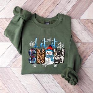 Op de afbeelding: Een groen sweatshirt met een sneeuwpop-afbeelding en de tekst "Let it Snow". De sneeuwpop draagt een rode muts en een blauwe sjaal. De tekst is omringd door sneeuwvlokken.