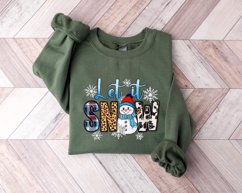 Op de afbeelding: Een groen sweatshirt met een sneeuwpop-afbeelding en de tekst "Let it Snow". De sneeuwpop draagt een rode muts en een blauwe sjaal. De tekst is omringd door sneeuwvlokken.