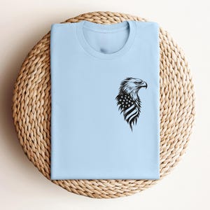 May include: A light blue t-shirt with a black and white graphic of an eagle's head with an American flag on its chest.