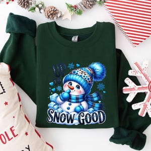 Peut inclure: Sweat-shirt vert avec un motif de bonhomme de neige et le texte "Up to Snow Good". Le bonhomme de neige porte un chapeau et une &eacute;charpe bleus et blancs.