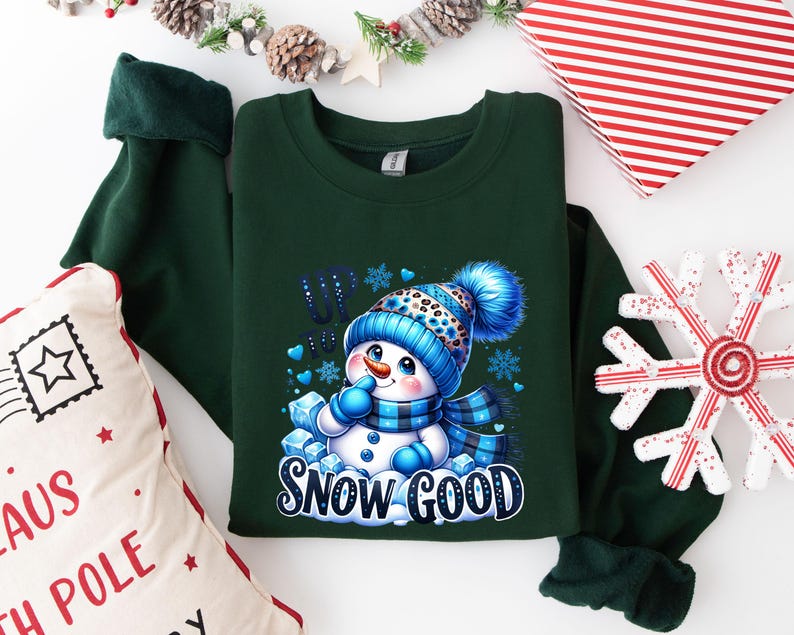 Peut inclure: Sweat-shirt vert avec un motif de bonhomme de neige et le texte "Up to Snow Good". Le bonhomme de neige porte un chapeau et une &eacute;charpe bleus et blancs.