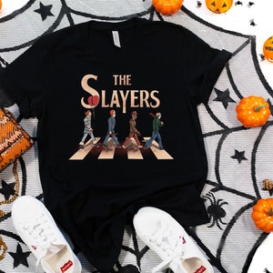 May include: Black t-shirt with a graphic of four figures walking across a crosswalk, with the text "The Slayers" above them. The figures are dressed in costumes reminiscent of horror movie characters. The background is a black and white spiderweb pattern.