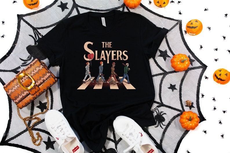 May include: Black t-shirt with a graphic of four figures walking across a crosswalk, with the text "The Slayers" above them. The figures are dressed in costumes reminiscent of horror movie characters. The background is a black and white spiderweb pattern.