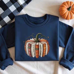 May include: A navy blue crewneck sweatshirt featuring a central graphic of a pumpkin constructed from open books with colorful spines and a green stem. The design displays various book pages and detailed bindings arranged in a circular shape.