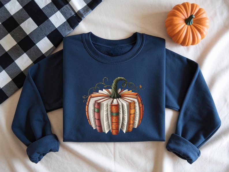 May include: A navy blue crewneck sweatshirt featuring a central graphic of a pumpkin constructed from open books with colorful spines and a green stem. The design displays various book pages and detailed bindings arranged in a circular shape.