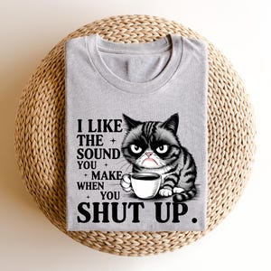May include: A gray t-shirt featuring a grumpy cartoon cat holding a coffee cup. The shirt has the text "I LIKE THE SOUND YOU MAKE WHEN YOU SHUT UP" in black font. The shirt is folded and placed on a woven, circular surface.