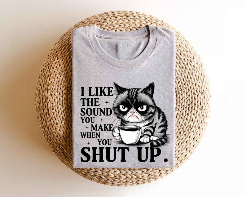 May include: A gray t-shirt featuring a grumpy cartoon cat holding a coffee cup. The shirt has the text "I LIKE THE SOUND YOU MAKE WHEN YOU SHUT UP" in black font. The shirt is folded and placed on a woven, circular surface.