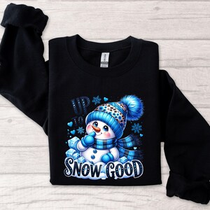 Peut inclure: Sweat-shirt noir avec un motif de bonhomme de neige bleu et blanc. Le bonhomme de neige porte un chapeau et une &eacute;charpe bleus et est entour&eacute; de flocons de neige bleus. Le texte "Up to Snow Good" est imprim&eacute; sous le bonhomme de neige.