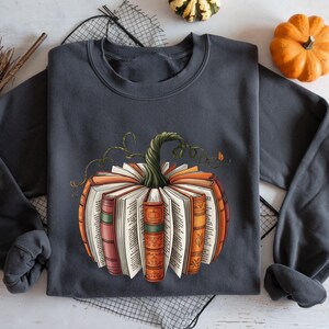 May include: A charcoal gray crewneck sweatshirt featuring a graphic print of a pumpkin constructed from open books. The books have colorful spines in shades of orange, red, and green, with a green, twisted vine stem emerging from the top center.