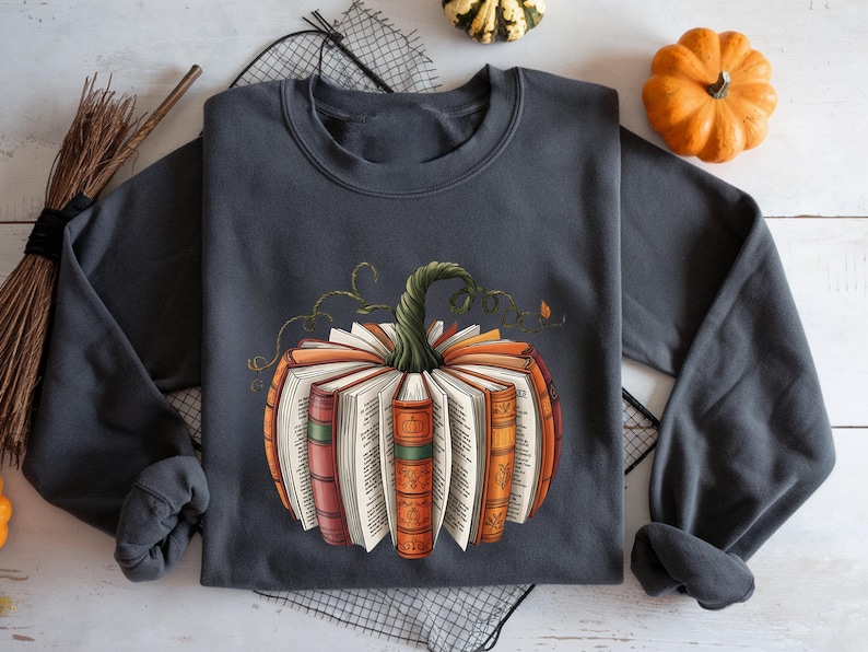 May include: A charcoal gray crewneck sweatshirt featuring a graphic print of a pumpkin constructed from open books. The books have colorful spines in shades of orange, red, and green, with a green, twisted vine stem emerging from the top center.