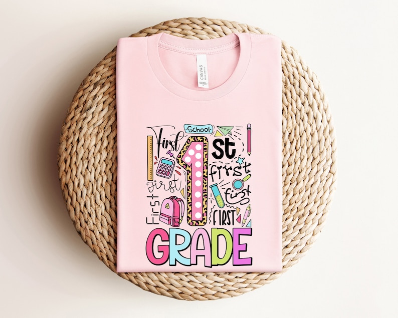 May include: A light pink t-shirt with a colorful graphic design for the first grade. The design includes the number "1" with a leopard print pattern, school supplies, and the words "First Grade". The shirt is folded on a woven surface.