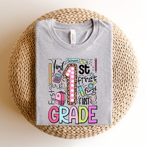 May include: A heather gray t-shirt with a colorful graphic design for first grade. The design includes the number 1 with a leopard print pattern, the words "First Grade", and school-themed illustrations like a ruler, pencil, and backpack.