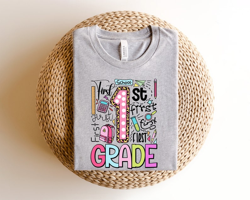 May include: A heather gray t-shirt with a colorful graphic design for first grade. The design includes the number 1 with a leopard print pattern, the words "First Grade", and school-themed illustrations like a ruler, pencil, and backpack.