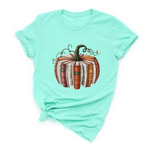 May include: A mint green short-sleeve t-shirt featuring a central graphic of a pumpkin constructed from open books. The pumpkin has a dark green stem with curling vines and pages fanned out to form the round shape of the gourd.