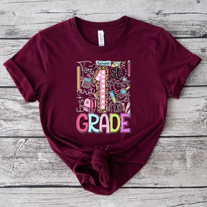 May include: Burgundy t-shirt with a colorful graphic for 1st grade. The design includes a large number 1 with a leopard print pattern, school supplies, and the words "1st Grade" in bright colors. The shirt is made of soft material.
