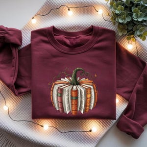 May include: A maroon long-sleeve sweatshirt featuring a central graphic of a pumpkin constructed from the pages and spines of various books. The pumpkin has a green stem and is rendered in shades of orange, brown, and cream, centered on the chest.