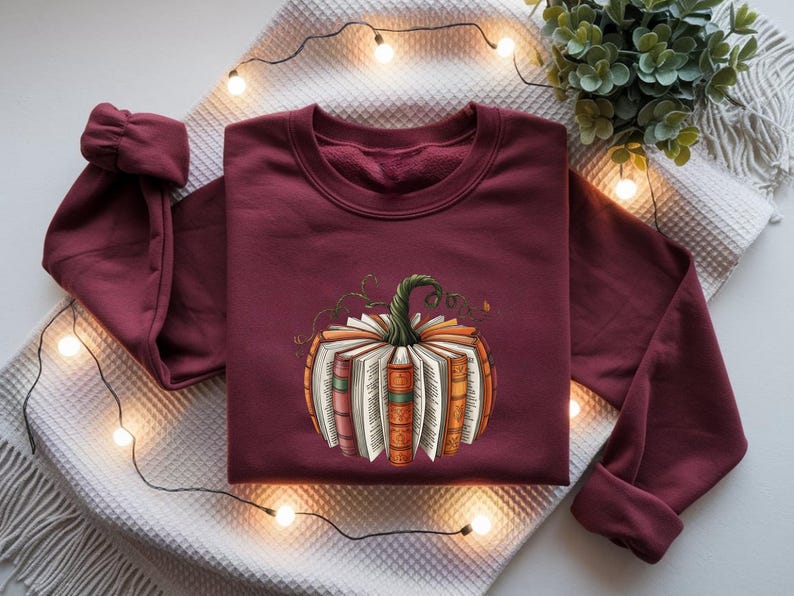 May include: A maroon long-sleeve sweatshirt featuring a central graphic of a pumpkin constructed from the pages and spines of various books. The pumpkin has a green stem and is rendered in shades of orange, brown, and cream, centered on the chest.