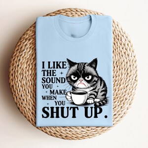 May include: Light blue t-shirt featuring a grumpy cat illustration holding a coffee cup. The shirt has the text "I LIKE THE SOUND YOU MAKE WHEN YOU SHUT UP." The cat is black and gray with a grumpy expression.