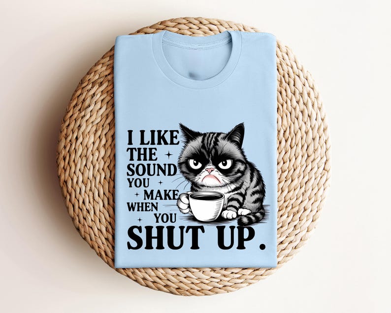May include: Light blue t-shirt featuring a grumpy cat illustration holding a coffee cup. The shirt has the text "I LIKE THE SOUND YOU MAKE WHEN YOU SHUT UP." The cat is black and gray with a grumpy expression.