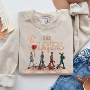 May include: A beige sweatshirt with a graphic print of horror movie characters walking across a crosswalk. The text "The Slayers" is printed above the characters.